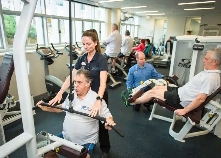 A physical therapist assists an older man on a lat‑pulldown machine while other people exercise on cardio and weight machines in a bright, busy gym. A physical therapist assists an older man on a lat‑pulldown machine while other people exercise on cardio and weight machines in a bright, busy gym.